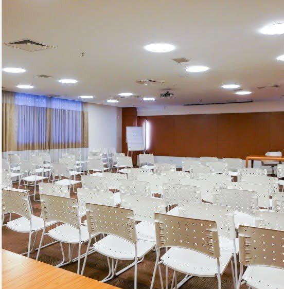Sala de treinamento com fileiras de cadeiras brancas, mesas de apoio, iluminação no teto e painel de madeira ao fundo.